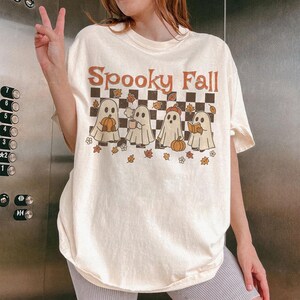 May include: Cream-colored t-shirt with the text "Spooky Fall" in brown and orange. The shirt features a checkered background with four cartoon ghosts holding a pumpkin, a mug, a pumpkin, and a book. Autumn leaves and flowers are scattered around the ghosts.