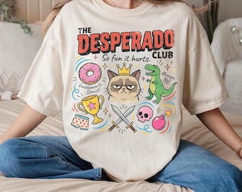 May include: Cream-colored t-shirt with a graphic design featuring the text "THE DESPERADO CLUB" and various cartoon illustrations. The design includes a donut, a grumpy cat wearing a crown, a green dinosaur, a trophy, crossed swords, a pink skull, and a bomb.