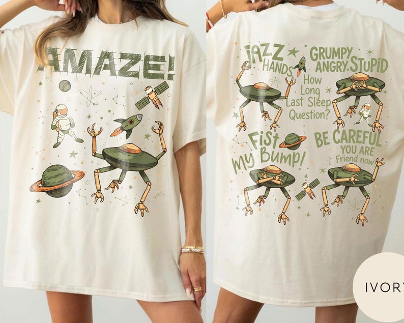 May include: Ivory-colored t-shirt with a whimsical space-themed design. The front features the word "AMAZE!" and illustrations of planets, a rocket, an astronaut, and a UFO. The back has text and more UFO illustrations.