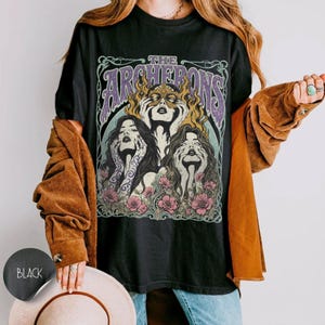 May include: A black t-shirt featuring a graphic print with the text THE ARCHERONS in purple letters. The design depicts three figures with their hands near their faces, surrounded by yellow flames and pink flowers, all framed by ornate lines.