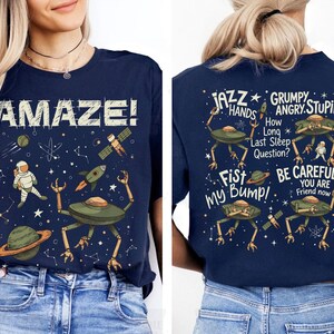 May include: Navy blue t-shirt with a space-themed graphic. The front features the word "AMAZE!" with an astronaut, planets, and rockets. The back displays alien figures and phrases like "JAZZ HANDS" and "BE CAREFUL".