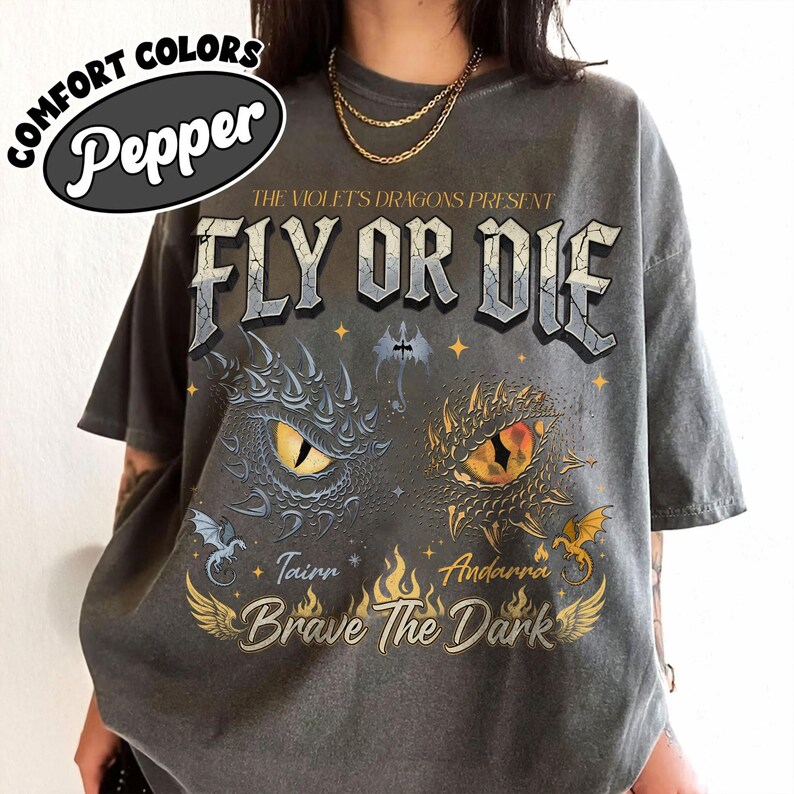 May include: Dark gray t-shirt with the text "FLY OR DIE" and dragon eye graphics. The shirt also features the text "THE VIOLET'S DRAGONS PRESENT", "Tainn", "Andarra", and "Brave The Dark". The shirt has a comfort colors label.