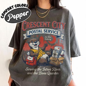 May include: Gray t-shirt with a graphic of an otter in a postal uniform, red mail bag, and the text "Crescent City Postal Service". The design includes a cityscape, moon, and the words "On Time Delivery". The shirt also says "Serving the Istros River and the Bone Quarter".