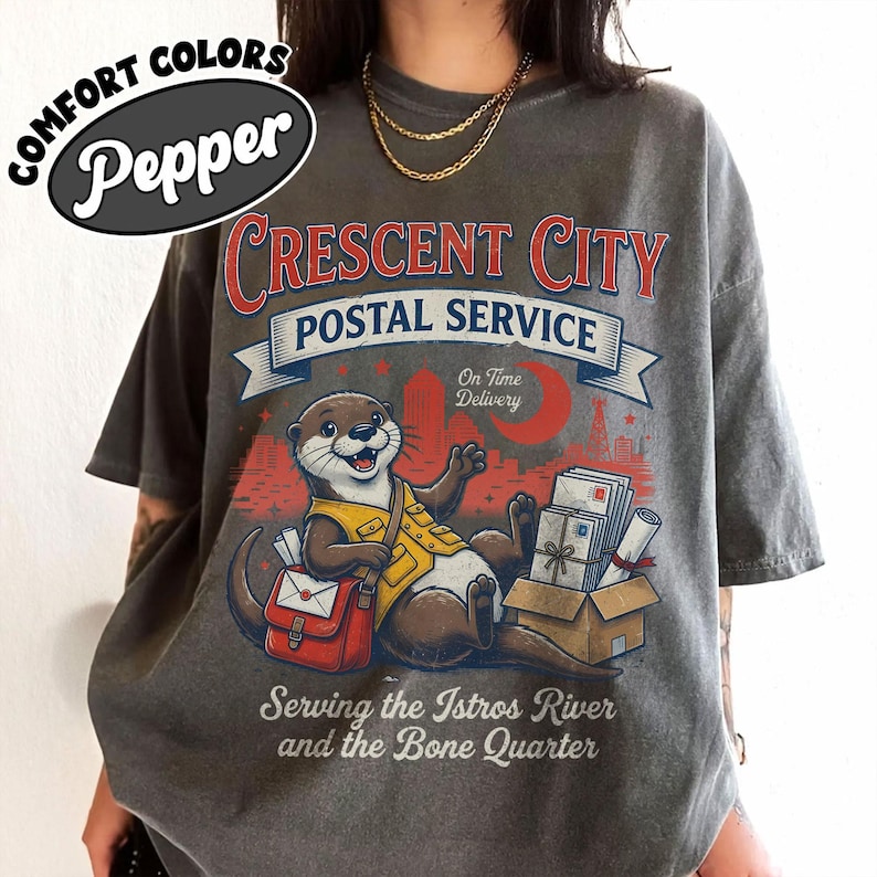 May include: Gray t-shirt with a graphic of an otter in a postal uniform, red mail bag, and the text "Crescent City Postal Service". The design includes a cityscape, moon, and the words "On Time Delivery". The shirt also says "Serving the Istros River and the Bone Quarter".