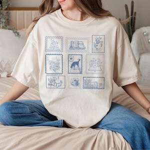 Puede incluir: Camiseta color crema con dise&ntilde;os de sellos postales azules. Cada sello muestra ilustraciones de libros, flores y un gato. La camiseta tiene un corte holgado y se usa con vaqueros azules.