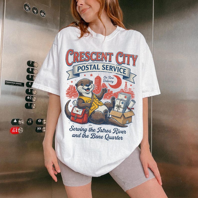 May include: White t-shirt featuring a cartoon otter in a postal worker uniform, with the text "Crescent City Postal Service." The design includes a red mail bag, a box of packages, and a cityscape. The shirt also has the text "Serving the Istros River and the Bone Quarter."