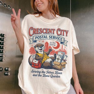 May include: Cream-colored t-shirt featuring a cartoon otter in a postal worker uniform. The shirt has the text "Crescent City Postal Service" above the otter, with additional text below that reads "Serving the Istros River and the Bone Quarter."