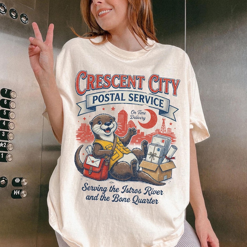 May include: Cream-colored t-shirt featuring a cartoon otter in a postal worker uniform. The shirt has the text "Crescent City Postal Service" above the otter, with additional text below that reads "Serving the Istros River and the Bone Quarter."