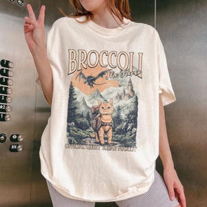 May include: A light beige t-shirt with a graphic print of a cat wearing a backpack and a dragon. The text on the shirt reads "Broccoli The Fierce Official Quest Squad Mascot".
