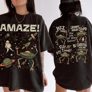May include: Black t-shirt with a space-themed graphic. The front features the word "AMAZE!" and illustrations of an astronaut, planets, a rocket, and a UFO. The back displays text and more UFO illustrations.