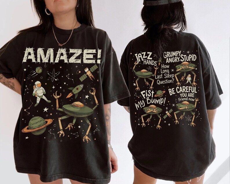 May include: Black t-shirt with a space-themed graphic. The front features the word "AMAZE!" and illustrations of an astronaut, planets, a rocket, and a UFO. The back displays text and more UFO illustrations.