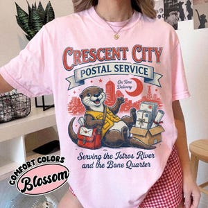 May include: Pink t-shirt with a graphic of an otter in a postal worker uniform, the text "Crescent City Postal Service", and the words "Serving the Istros River and the Bone Quarter". The shirt also has the word "Blossom".