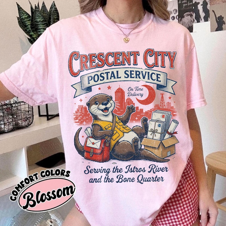 May include: Pink t-shirt with a graphic of an otter in a postal worker uniform, the text "Crescent City Postal Service", and the words "Serving the Istros River and the Bone Quarter". The shirt also has the word "Blossom".