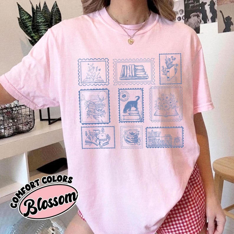 Puede incluir: Camiseta rosa claro con dise&ntilde;os de sellos postales azules que representan libros, flores y un gato. El texto "Comfort Colors Blossom" est&aacute; impreso en la parte inferior izquierda. Prenda casual y c&oacute;moda.