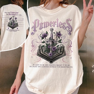 May include: A white t-shirt with a purple and black graphic design of a book with a sword and flowers. The text "Powerless" is printed in a gothic font above the graphic. The text "If I am to be her enemy, I want it to be because she loathes herself for wanting me" is printed below the graphic.