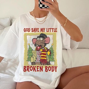 May include: White t-shirt featuring a cartoon rat character wearing a hat, scarf, and jacket, with the text "GOD SAVE MY LITTLE BROKEN BODY" in red and black. The graphic is framed by a yellow border with Christmas trees.