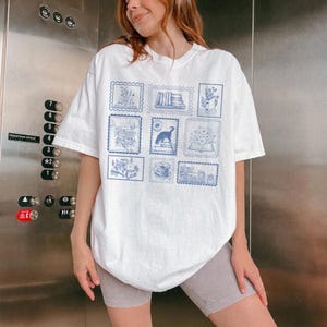Puede incluir: Camiseta blanca con dise&ntilde;os de sellos postales azules que representan libros, flores y un perro. La camiseta tiene un corte holgado y de gran tama&ntilde;o.