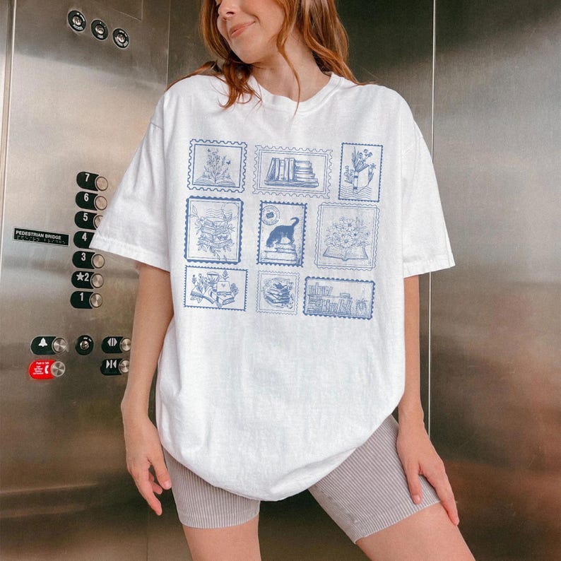 Puede incluir: Camiseta blanca con dise&ntilde;os de sellos postales azules que representan libros, flores y un perro. La camiseta tiene un corte holgado y de gran tama&ntilde;o.