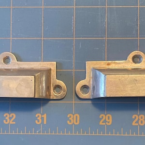 May include: Two antique metal drawer pulls. Each pull is rectangular with a recessed handle area and rounded corners. They have a silver finish and are shown against a blue grid background. Each pull measures approximately 4 inches long.