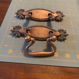 May include: Two antique bronze drawer pulls. Each pull has an oval center with decorative flourishes on the ends and a curved handle. The pulls are set against a green cutting mat with measurement markings.