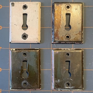 Puede incluir: Cuatro cubiertas de cerradura rectangulares antiguas en tonos de blanco roto, marrón y negro. Cada cubierta tiene una forma de cerradura en el centro y dos agujeros circulares. Las cubiertas muestran signos de desgaste y pintura descascarillada.