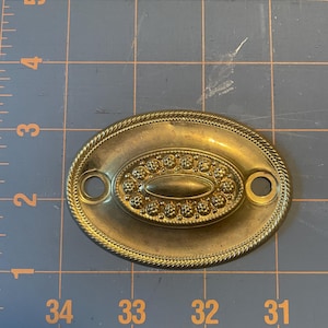 Puede incluir: Tirador de cajón ovalado de latón con un borde decorativo en forma de cuerda. El centro presenta un diseño ovalado en relieve con pequeños adornos circulares. Hay dos agujeros para el montaje. Una pieza de ferretería de estilo vintage.