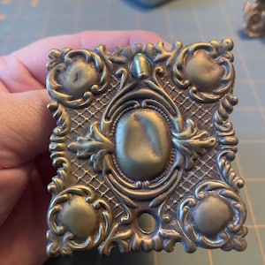 Könnte beinhalten: Verziertes Möbelbeschlag in Messingoptik. Das quadratische Stück hat eine ovale Mitte und vier abgerundete Ecken, die von detaillierten Schnörkeln und einem rautenförmigen Hintergrund umgeben sind. Unten ist ein kleines Loch sichtbar. Für Schränke oder Schubladen.