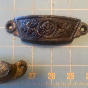 Op de afbeelding: Close-up van antieke metalen lade grepen. De grepen zijn donkergrijs met sierlijke bloemmotieven. De grepen hebben twee gaten voor montage. De achtergrond is een blauwe snijmat met meetmarkeringen.