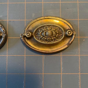 Puede incluir: Un tirador de cajón ovalado de latón antiguo con un diseño floral en el centro. El tirador tiene un patrón en relieve detallado y pequeños acentos circulares en cada extremo. El herraje es probablemente para un armario o una cómoda.