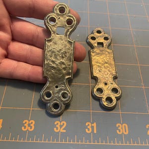 May include: Two antique brass-toned metal decorative pulls. Each pull has an ornate design with a rectangular center and decorative top and bottom. The pulls have a hammered texture and are of different sizes.