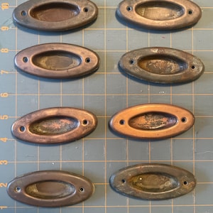 Puede incluir: Ocho tiradores de cajón ovalados de bronce antiguo, cada uno con dos agujeros. Los tiradores tienen un centro empotrado y una pátina desgastada, que muestra signos de edad y uso. Están dispuestos sobre un fondo de cuadrícula, lo que sugiere un taller o un entorno artesanal.