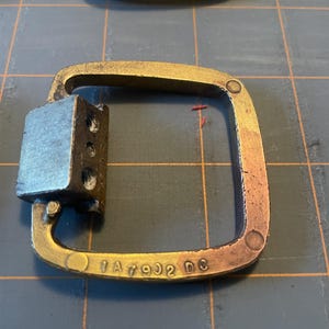 May include: Close-up of a brass-colored metal buckle with a square shape and a rectangular attachment. The buckle has the stamped text "1A7902 D3". The metal has a slightly aged appearance.