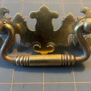 May include: Vintage brass drawer pull with an elaborate, decorative design. It features a central floral pattern and curved handle supports. The handle is a cylindrical bar with ribbed detailing. This hardware is likely for a cabinet or dresser.