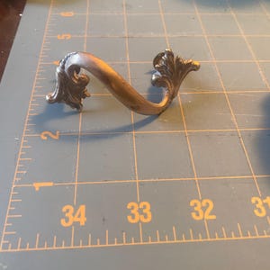 Może przedstawiać: Antyczny mosiężny uchwyt do szuflady o ozdobnym, zakrzywionym kształcie. Uchwyt posiada dekoracyjne kwiatowe akcenty na każdym końcu i delikatnie łukowaty uchwyt. Mosiądz ma ciepłą, postarzaną patynę.