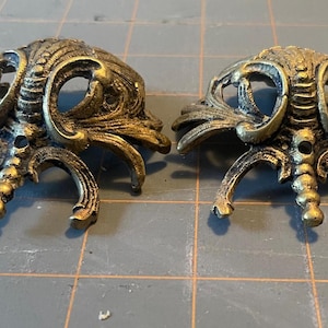 Peut inclure: Deux ferrures de meubles ornées de style antique. Chaque pièce est en métal de couleur bronze avec des motifs tourbillonnants complexes et des pieds incurvés. La quincaillerie a un aspect vieilli.