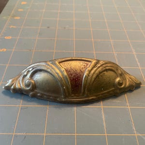 Może przedstawiać: Antyczny uchwyt do szuflady w odcieniu mosiądzu o ozdobnym wzorze. Uchwyt ma zakrzywiony kształt z dekoracyjnymi zawijasami na końcach i czerwonobrązowym środkiem. Okucia są prawdopodobnie do szafki lub komody.