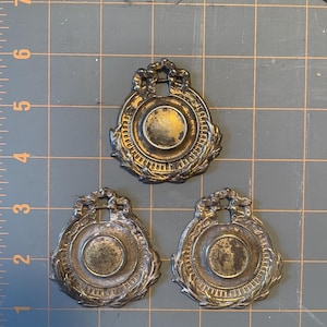 Op de afbeelding: Drie antieke, aangetaste metalen medaillons met een cirkelvormig ontwerp en laurierkransdetails. Elk medaillon heeft een lus aan de bovenkant om op te hangen. De medaillons liggen op een grijs rasteroppervlak.