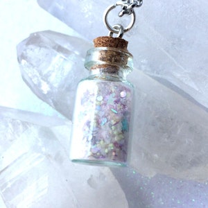 May include: A silver chain necklace with a small glass bottle pendant filled with iridescent glitter. The bottle has a cork stopper and is hanging against a background of clear quartz crystals.