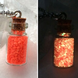 May include: A glass bottle necklace with a cork stopper filled with orange glow-in-the-dark sand. The necklace is shown in two images, one in daylight and one in the dark.