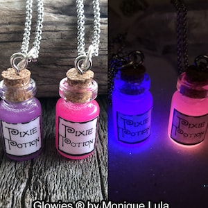 Könnte beinhalten: Zwei Glasflaschen mit Korkstopfen und Etiketten mit der Aufschrift "Pixie Potion". Die Flaschen sind mit einer glitzernden Flüssigkeit gefüllt und an silbernen Ketten befestigt. Die Flaschen leuchten im Dunkeln, eine in Lila und die andere in Pink.