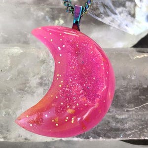 May include: A pink crescent moon pendant necklace with glitter accents. The pendant hangs from a rainbow-colored chain. The necklace is displayed on clear quartz crystals. The text "GLOWIES by MONIQUE LULA" is visible.