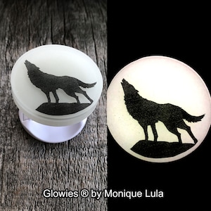 May include: A white glow-in-the-dark round disc with a black silhouette of a wolf howling at the moon. The disc is on a wooden surface. "Glowies® by Monique Lula" is written below the disc.