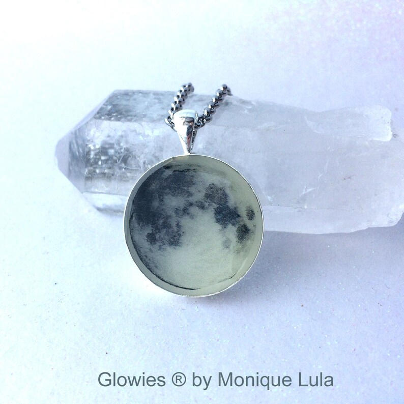 Puede incluir: Un collar de plata con un colgante redondo que presenta una imagen en blanco y negro de la luna. El colgante est&aacute; sobre una superficie blanca.