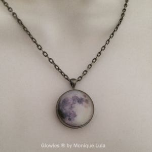 Puede incluir: Un collar de cadena plateada con un colgante redondo que presenta una imagen de primer plano de la luna. La luna es de color gris claro con cr&aacute;teres grises oscuros y un brillo blanco alrededor de los bordes. El texto "Glowies &reg; by Monique Lula" est&aacute; impreso debajo del colgante.