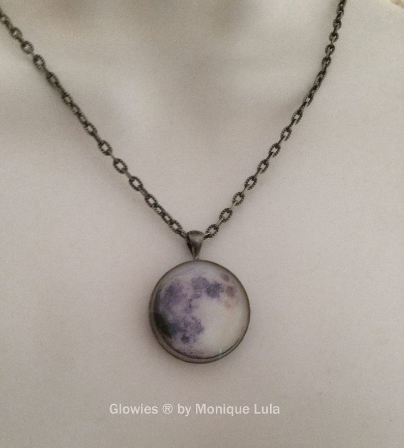 Puede incluir: Un collar de cadena plateada con un colgante redondo que presenta una imagen de primer plano de la luna. La luna es de color gris claro con cr&aacute;teres grises oscuros y un brillo blanco alrededor de los bordes. El texto "Glowies &reg; by Monique Lula" est&aacute; impreso debajo del colgante.