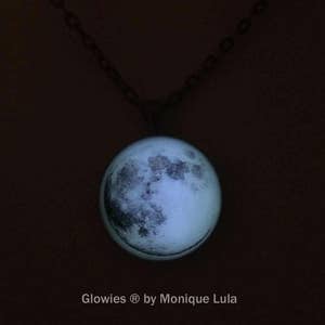 Puede incluir: Un collar con cadena plateada y un colgante redondo de vidrio con una imagen de la luna que brilla en la oscuridad. El texto "Glowies &reg; by Monique Lula" est&aacute; impreso debajo del colgante.