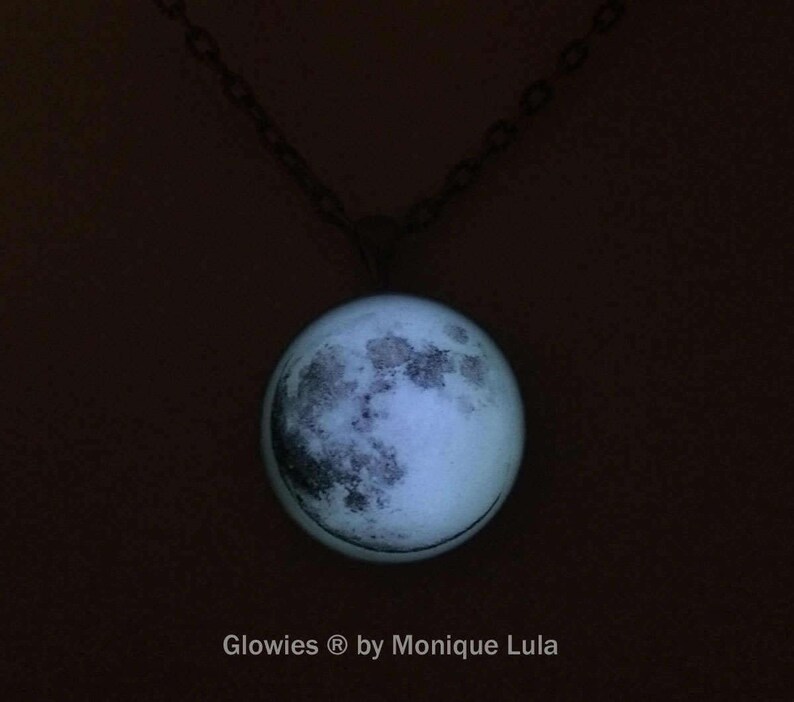 Puede incluir: Un collar con cadena plateada y un colgante redondo de vidrio con una imagen de la luna que brilla en la oscuridad. El texto "Glowies &reg; by Monique Lula" est&aacute; impreso debajo del colgante.