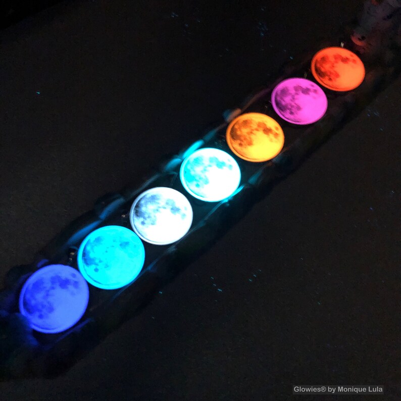 Puede incluir: Una fila de siete llaveros con forma de luna que brillan en la oscuridad, en diferentes colores, incluyendo azul, azul verdoso, blanco, naranja, rosa y rojo. Los llaveros est&aacute;n dispuestos en l&iacute;nea sobre una superficie oscura. Glowies&reg; by Monique Lula.