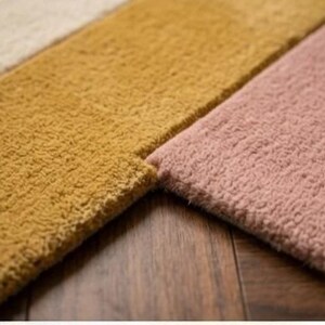 May include: Close-up of three sections of a plush rug in varying colors. The sections are a light beige, a mustard yellow, and a dusty rose pink. The rug has a textured, looped pile. The rug is placed on a dark wood surface.