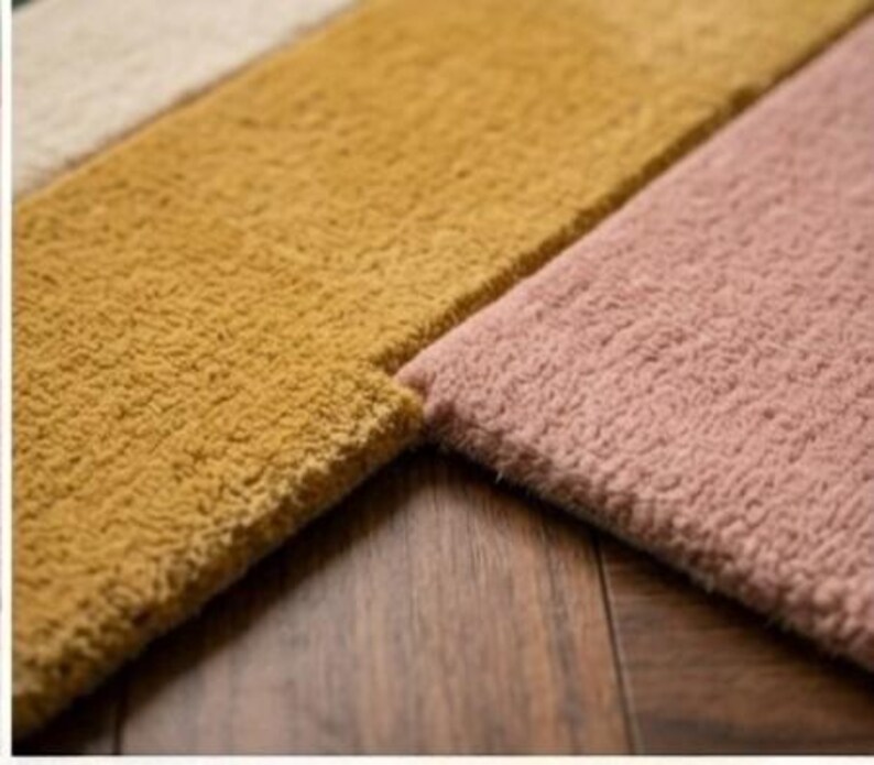 May include: Close-up of three sections of a plush rug in varying colors. The sections are a light beige, a mustard yellow, and a dusty rose pink. The rug has a textured, looped pile. The rug is placed on a dark wood surface.